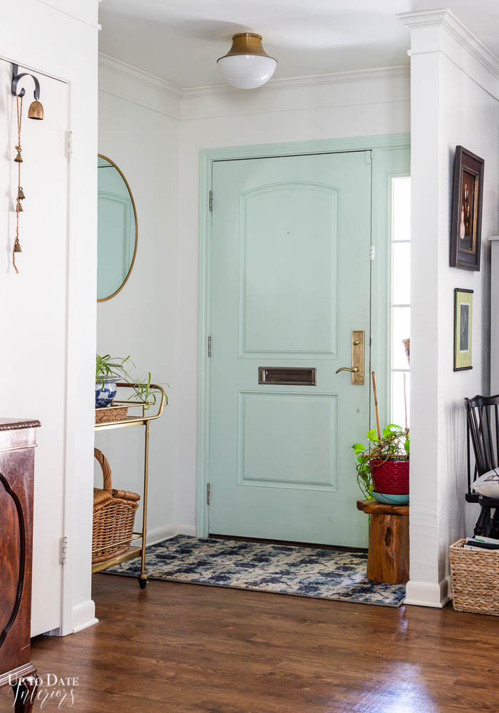 A cute green front door in a white foyer with plants, gold mirror, art, rug, and wood floors.  Beautiful brass hardware. 