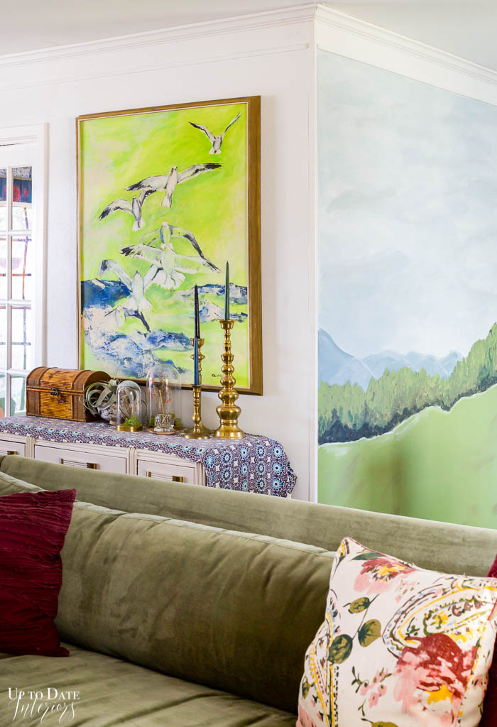 A colorful and eclectic open layout living areas showing a partial view of the dining room wall hand painted mural and bold bird art in yellow and blue.  A velvet sage green sofa with pillows. 