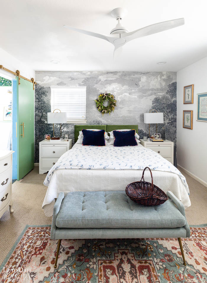 A master bedroom with a grey anthropologie landscape mural on the wall and a dried fern wreath hanging above a green velvet headboard wtih white bedding, white side tables and lamps, and dark blue velvet throw pillows. A light blue upholstered modern bench is at the foot of the bed on a traditional rug. To the left is a bold green barn door with gold hardware and pull leading to the ensuite bathroom. 