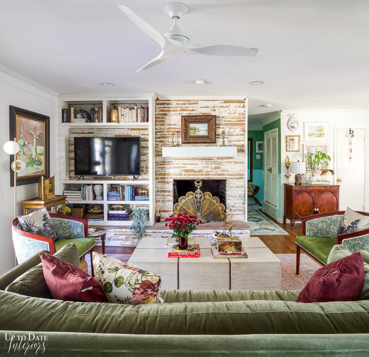 A view of the living area in an open layout with a german smear stone fireplace and a white built in bookcase decorated with books and a large tv.  A green velvet sofa, white and chrome coffee table and a pair of green floral barrel chairs on either side make a cozy seating arrangement. 
