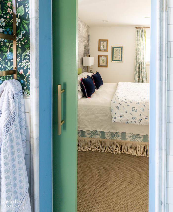 A view from the bathroom showing an opened green sliding door with a brass pull and blue threshold and a glimpse of marble molding, white tile wainscoting and floral green wallpaper.  The view is into the bedroom with white walls and blue and white textiles and neutral landscape mural. 