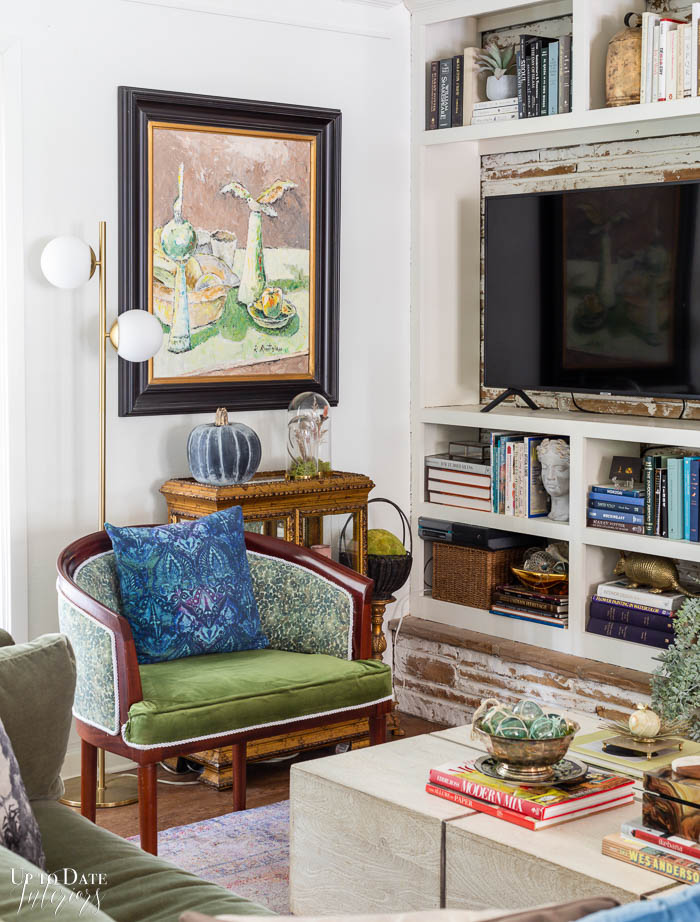 A corner of a white and green living room featuring large art, bulit in bookcase, and colorful and eclectic furniture and decor.  A heirloom pumpkin is displayed for Fall. 
