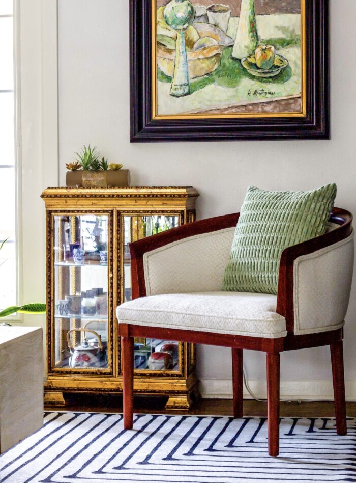 A reupholstered white and wood barrel chair sits in the corner with a striped rug and gold display cabinet with green art. 