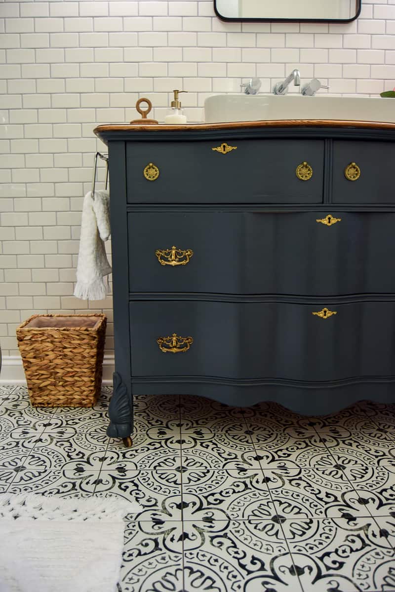 Patterned tile floor in a bathroom with dark vanity from dresser, and white subway tile wall. 