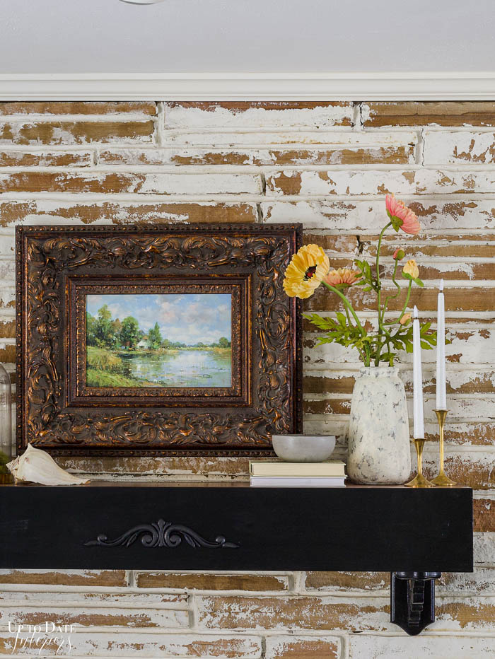 A close up view of artificial flower bouquet of poppies look beautiful in a stone vase on a black mantel with a landscape oil painting, candles, and german smear fireplace surround. 